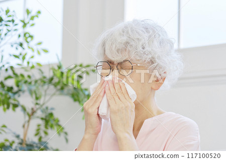 Senior woman blowing her nose in living room 117100520