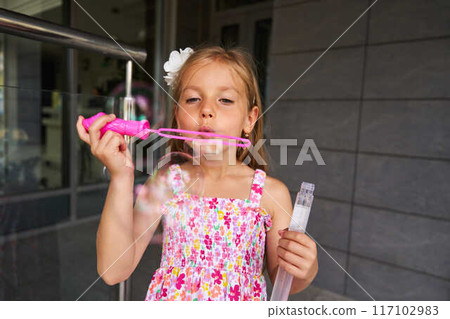 Young Girl Blowing Colorful Bubbles While Wearing Floral Dress Outside a Modern Building 117102983