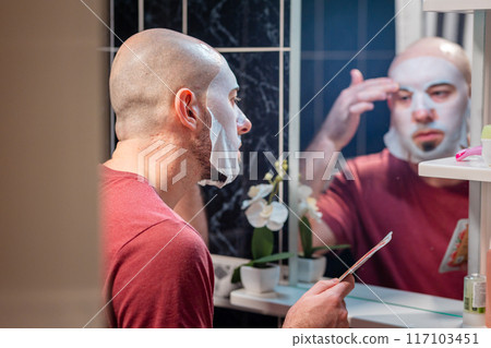 View from door at young adult Caucasian bald bearded man using cloth mask to moisturize the skin of his face. Beauty routine and skin care View from door at young adult Caucasian bald bearded man using cloth mask to moisturize the skin of his face. Beauty routine and skin care 117103451