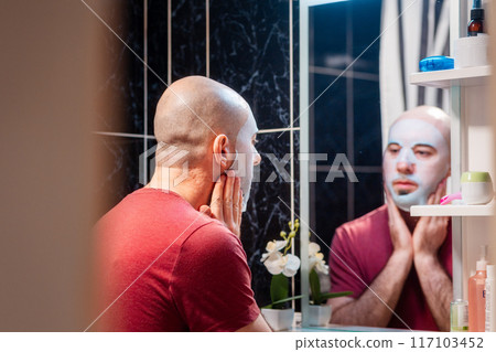 Back view from door of young adult Caucasian bald bearded man using cloth mask to moisturize the skin of his face. Beauty routine and skin care concept 117103452
