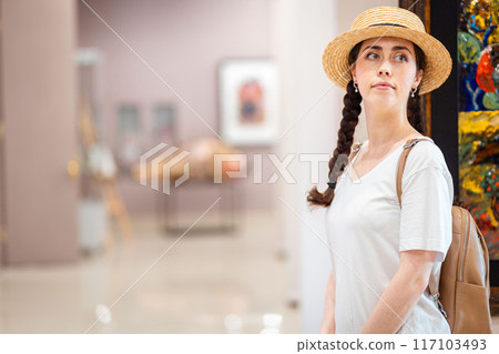 Historic education. Portrait of pretty young Caucasian woman wearing straw hat admiring paintings in the gallery. Copy space. Concept of visit museum and cultural education 117103493