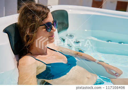 Woman wearing sunglasses and swimsuit relaxing in outdoor hot tub with bubbling water and enjoying sunny day. Female resting in outdoor jacuzzi with beautiful natural landscape Woman wearing sunglasses and swimsuit relaxing in outdoor hot tub with bubbling water and enjoying sunny day. Female resting in outdoor jacuzzi with beautiful natural landscape 117108406