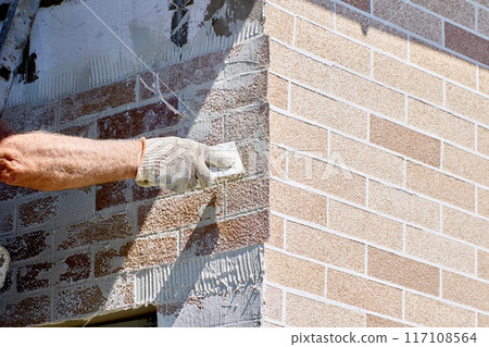 Installing flexible thin panel of fake brick on facade of building, construction worker removes excess adhesive mortar with steel putty knife. 117108564
