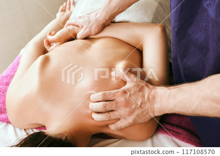 Shoulder area, upper back and neck deep stroking and pressure tissue massage, young white woman receiving physiotherapy while lying face down on massage table. Shoulder area, upper back and neck deep stroking and pressure tissue massage, young white woman receiving physiotherapy while lying face down on massage table. 117108570