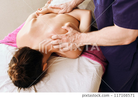 Caucasian woman receives deep tissue massage on her back and neck while lying face down on massage table. 117108571