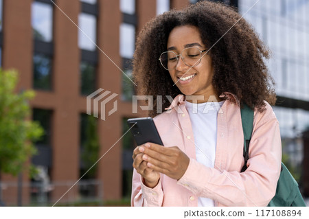 African American woman student with glasses and backpack smiling while using smartphone outdoors in urban setting. Confident, connected, and engaged in a city environment. African American woman student with glasses and backpack smiling while using smartphone outdoors in urban setting. Confident, connected, and engaged in a city environment. 117108894