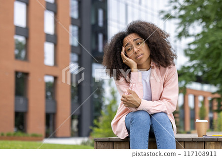Pensive young woman with curly hair sitting outdoors in urban environment. She appears thoughtful and contemplative. Glasses add intellectual look. Modern building background suggests urban lifestyle Pensive young woman with curly hair sitting outdoors in urban environment. She appears thoughtful and contemplative. Glasses add intellectual look. Modern building background suggests urban lifestyle 117108911