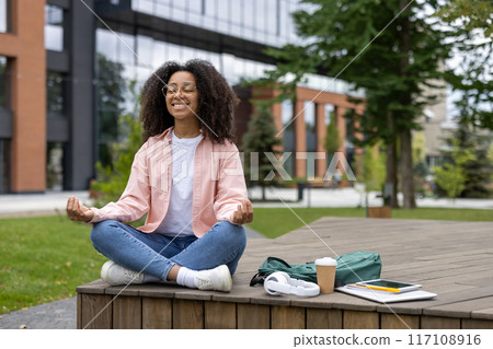 Young African American woman meditating outdoors, sitting cross-legged on wooden platform in urban park. Relaxing student in pink shirt and jeans, with backpack, coffee cup, and tablet beside her. 117108916