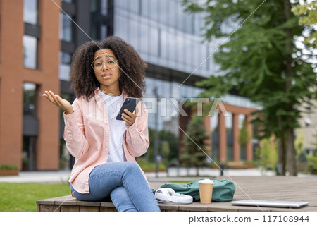 African american woman student looking confused while using smartphone outdoors on campus. Seen wearing casual clothes with laptop, coffee cup, and backpack nearby. Concept of confusion, campus life African american woman student looking confused while using smartphone outdoors on campus. Seen wearing casual clothes with laptop, coffee cup, and backpack nearby. Concept of confusion, campus life 117108944