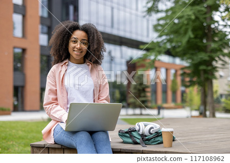 Young african american woman student sitting outdoors on university campus, working on laptop. Relaxed and focused, enjoying study time. Concept of education, technology, youth, and college life. Young african american woman student sitting outdoors on university campus, working on laptop. Relaxed and focused, enjoying study time. Concept of education, technology, youth, and college life. 117108962
