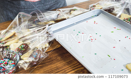 Carefully packaging Christmas cutout cookies, half-dipped in chocolate and presented in clear cellophane wrapping, perfect for festive gifting. 117109073
