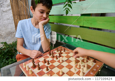 Boys playing chess outdoors in a garden setting on a wooden table 117109424