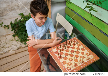 Young boy playing chess outside on a wooden patio during a sunny day 117109456