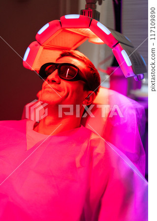 Handsome male with hairloss problem in protective goggles having LED light hair photodynamic therapy treatment after hair transplant at clinic. Man client having non-invasive type of phototherapy. Handsome male with hairloss problem in protective goggles having LED light hair photodynamic therapy treatment after hair transplant at clinic. Man client having non-invasive type of phototherapy. 117109890