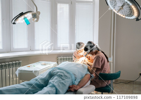 Remote view of two doctors collecting hair follicles from donor area of patient for hair transplantation. Process of hair transplant with FUE Sapphire method in clinic. Concept of baldness treatment Remote view of two doctors collecting hair follicles from donor area of patient for hair transplantation. Process of hair transplant with FUE Sapphire method in clinic. Concept of baldness treatment 117109898