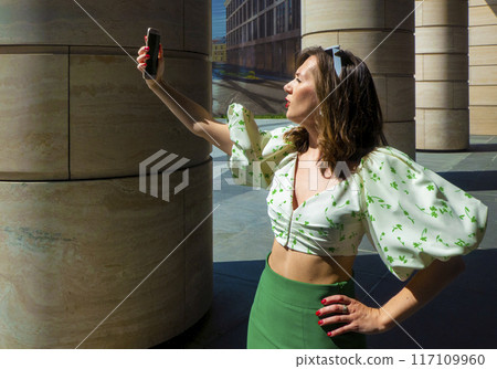Joyful young caucasian woman winks, bites her tongue, takes selfie on phone. Blonde model wears hat in sunny weather. Positive emotions concept Joyful young caucasian woman winks, bites her tongue, takes selfie on phone. Blonde model wears hat in sunny weather. Positive emotions concept 117109960