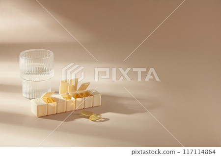Beige background photography for pharmaceutical product advertising, was taken from high angle. A yellow pill box containing vitamin or fish oil capsule tablets, copy space for adding text Beige background photography for pharmaceutical product advertising, was taken from high angle. A yellow pill box containing vitamin or fish oil capsule tablets, copy space for adding text 117114864