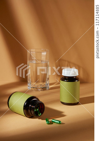 Brown background photography with medicine concept for advertisement from front view. Two brown supplement bottles with blank green label, featured on the table next to a glass of water 117114885