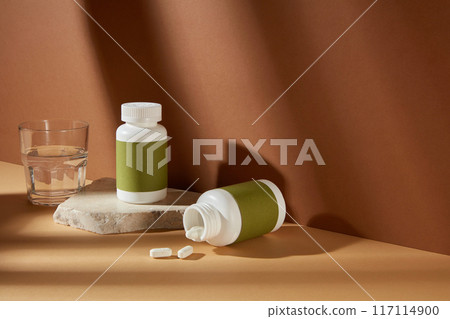 The empty green label pill bottle displayed on yellow countertop, over brown background with some scattered white pills and a glass of water. High angle shot photo, blank space for designing The empty green label pill bottle displayed on yellow countertop, over brown background with some scattered white pills and a glass of water. High angle shot photo, blank space for designing 117114900