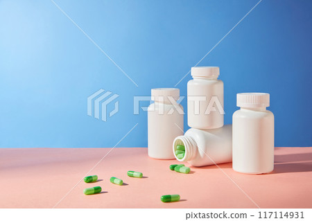 Blue and pink background photograph for medicine advertisement, a few white unlabeled medicine bottles displayed on the right and green capsules scattered flat lay on pink flat. Front view, copy space 117114931