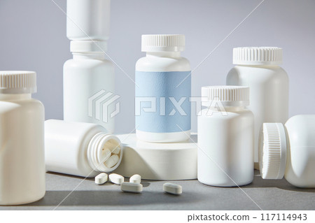 Advertisement pic with pharmaceutical theme from over white background. Plenty medicine jars without label displayed on the gray table, a blue label one stands out on the podium 117114943