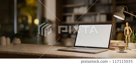 A laptop computer on a table in a modern room at night, illuminated by a table lamp. A laptop computer on a table in a modern room at night, illuminated by a table lamp. 117115035