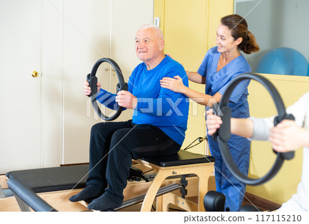 Healthcare professional helps an elderly man restore health with gymnastic ring in pilates class 117115210