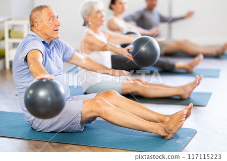 Group of mature people in sportswear exercising Pilates with soft balls while sitting on mats in fitness studio Group of mature people in sportswear exercising Pilates with soft balls while sitting on mats in fitness studio 117115223