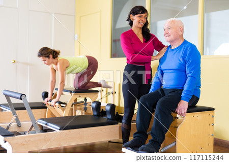 Elderly man practicing pilates on wunda chair under guidance of Hispanic female trainer 117115244
