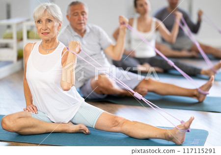 Elderly woman performs stretches elastic band with hand and foot while sitting on mat. 117115291