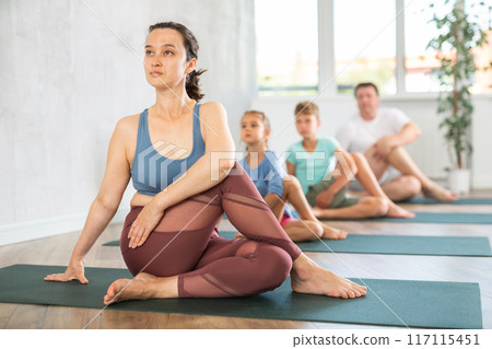 Middle-aged woman sitting in lord of fish pose, family in background Middle-aged woman sitting in lord of fish pose, family in background 117115451
