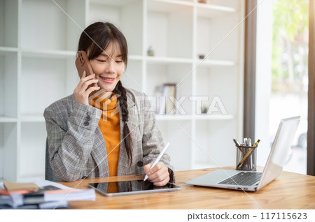 A busy young Asian businesswoman is talking on the phone while working on her digital tablet. A busy young Asian businesswoman is talking on the phone while working on her digital tablet. 117115623