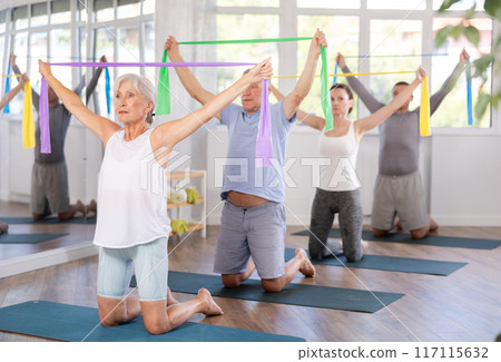 Elderly woman kneeling performs exercises with wide elastic band during Pilates workout. 117115632