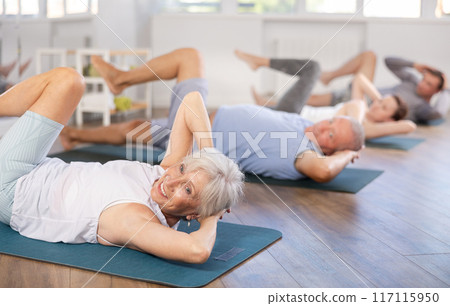 Elderly woman performs exercises to strengthen abdominal muscles during group workout 117115950