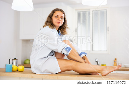 Lovely sexy girl in shirt posing while sitting on the table of modern kitchen Lovely sexy girl in shirt posing while sitting on the table of modern kitchen 117116072