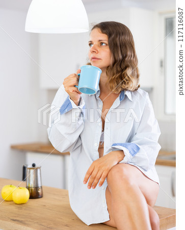 Woman sitting on kitchen countertop, drinking coffee Woman sitting on kitchen countertop, drinking coffee 117116087