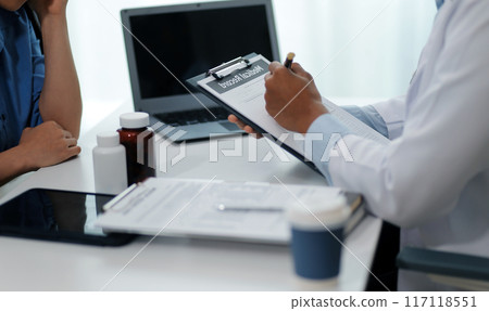 Doctor and patient sitting near each other at the wooden desk in clinic. Female physician's pointing to a records form. Medicine concept Doctor and patient sitting near each other at the wooden desk in clinic. Female physician's pointing to a records form. Medicine concept 117118551