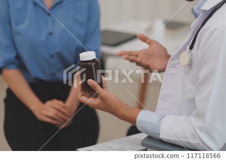 Confident doctor man holding a pill bottle and writing while talking with senior patient and reviewing his medication at office room. 117118566