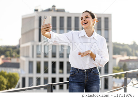 Smiling woman taking selfie on rooftop with cityscape background 117119763