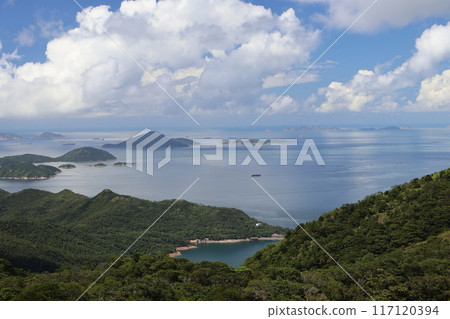 A view of the ocean beyond the mountains covered with green trees, countless islands and ships, the faintly visible opposite shore, white clouds and blue sky. A view of the ocean beyond the mountains covered with green trees, countless islands and ships, the faintly visible opposite shore, white clouds and blue sky. 117120394