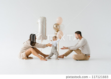 One year old baby takes the first steps from mom to dad against the background of festive birthday balloons 117121883