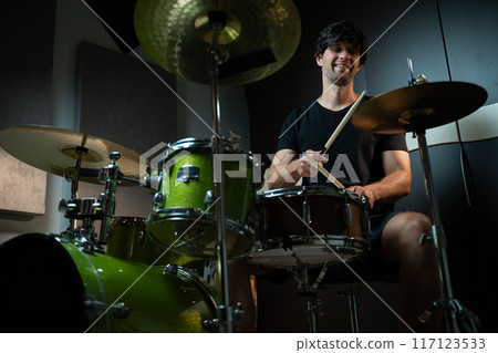 A man plays drums in a recording studio, seated behind a green drum set with black bass drum and two cymbals. 117123533