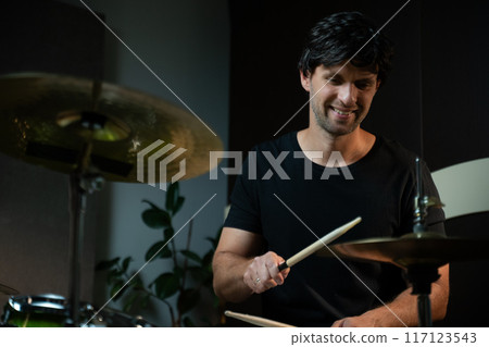 A man in a black shirt smiles while playing hi-hat with a drumstick. He is sitting behind a drum kit in a room to the left of which a plant is visible. 117123543