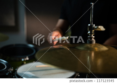 The drummer hits the hi-hat with a drumstick, making a loud, sonorous sound. The drummer is wearing a black shirt, and the cymbal is large and golden. The camera is aimed at the plate in close-up 117123545