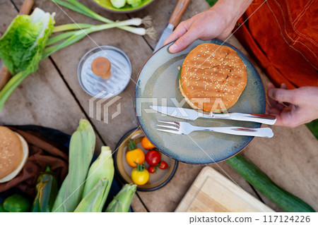Top view of man holding plate with homemade grilled hamburger and cutlery. Summer outdoor barbecue. 117124226