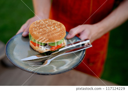 Close up of man holding plate with homemade grilled hamburger and cutlery. Summer outdoor barbecue. Close up of man holding plate with homemade grilled hamburger and cutlery. Summer outdoor barbecue. 117124229