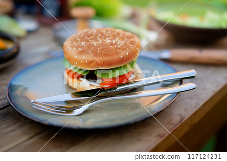 Close up of plate with homemade grilled hamburger and cutlery. Summer outdoor barbecue. 117124231