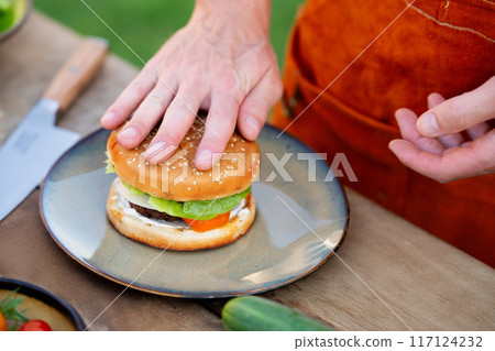 Close up of man assembling burgers with toppings, putting toasted bun on top of hamburger. Summer outdoor barbecue. 117124232