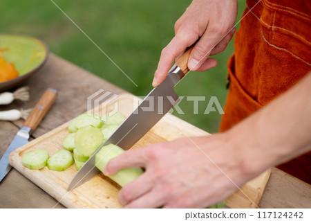 Close up of man holding sharp knife, chopping cucumber. Preparing vegetables for an outdoor barbecue. 117124242