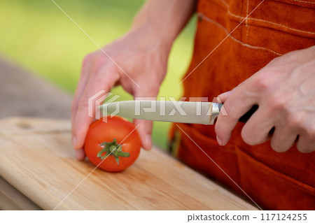 Close up of man holding sharp knife, slicing juicy red tomato. Preparing vegetables for an outdoor barbecue. 117124255